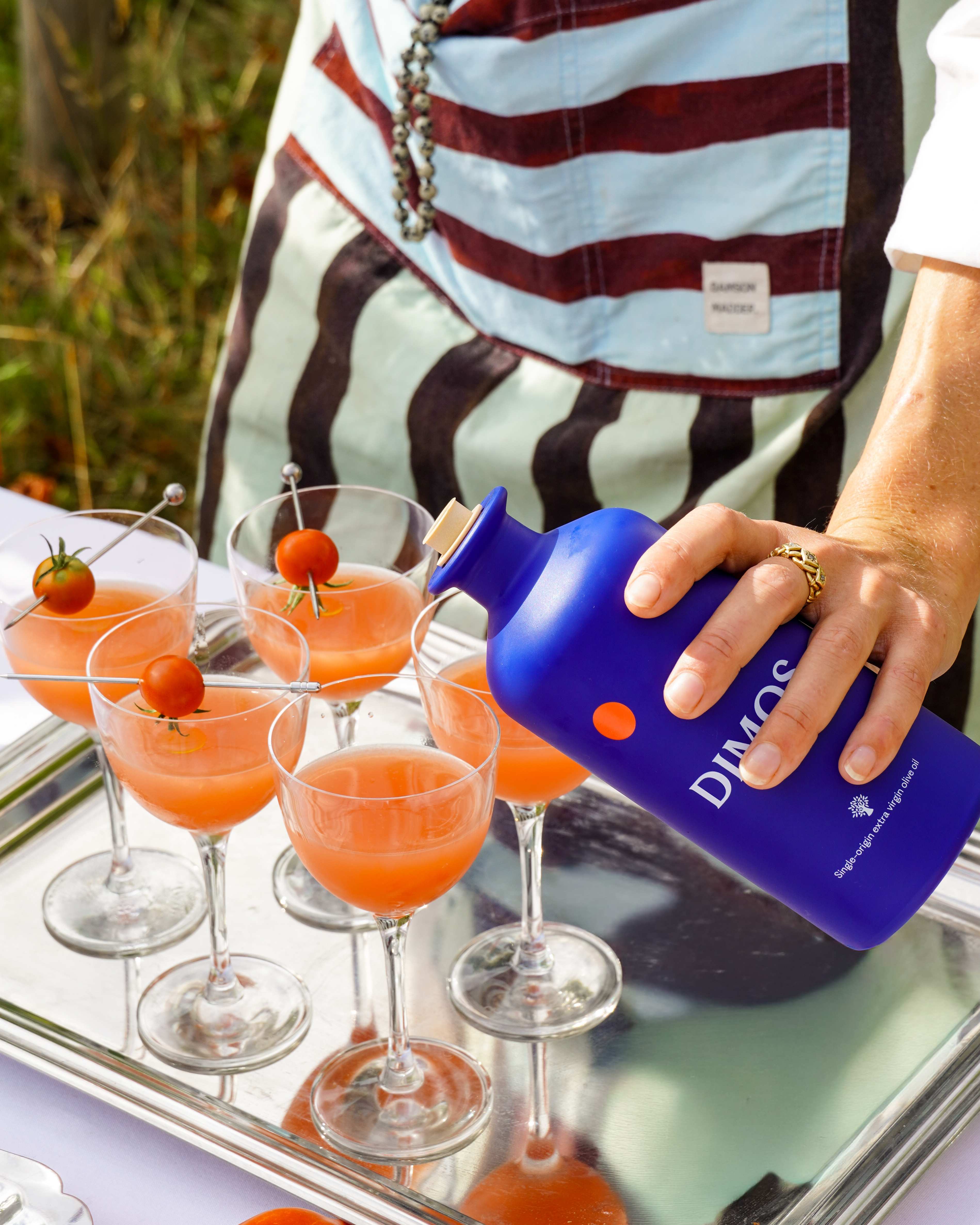 A hand pouring DIMOS olive oil from a blue bottle over a tray of tomato cocktails